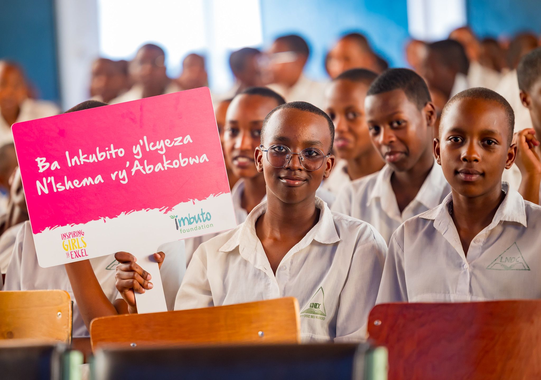 A student in glasses holds a pink 'Inkubito y'Icyeza' sign, classmates beside her