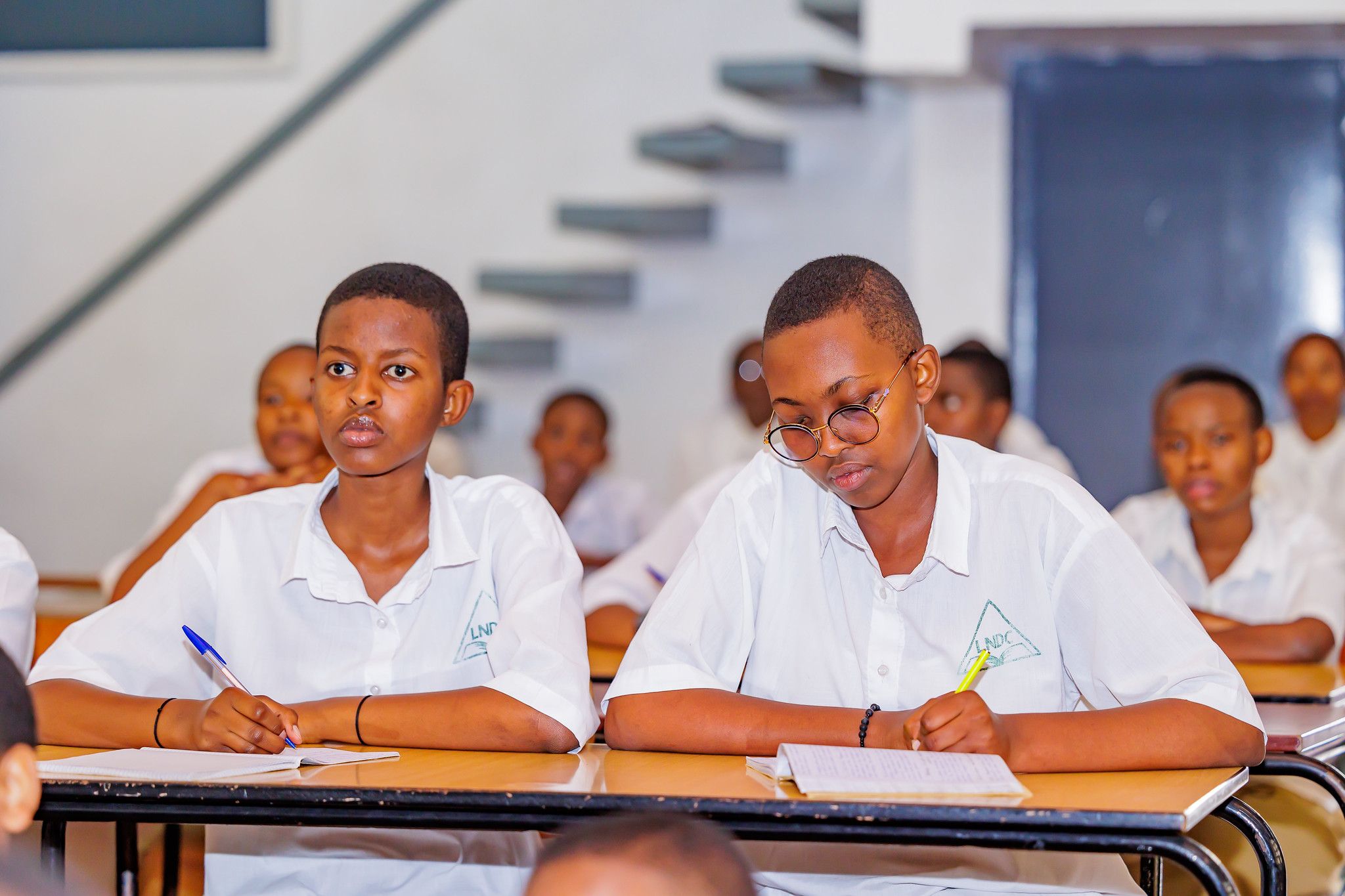 Two students at a shared desk, one writing in a notebook, the other listening closely