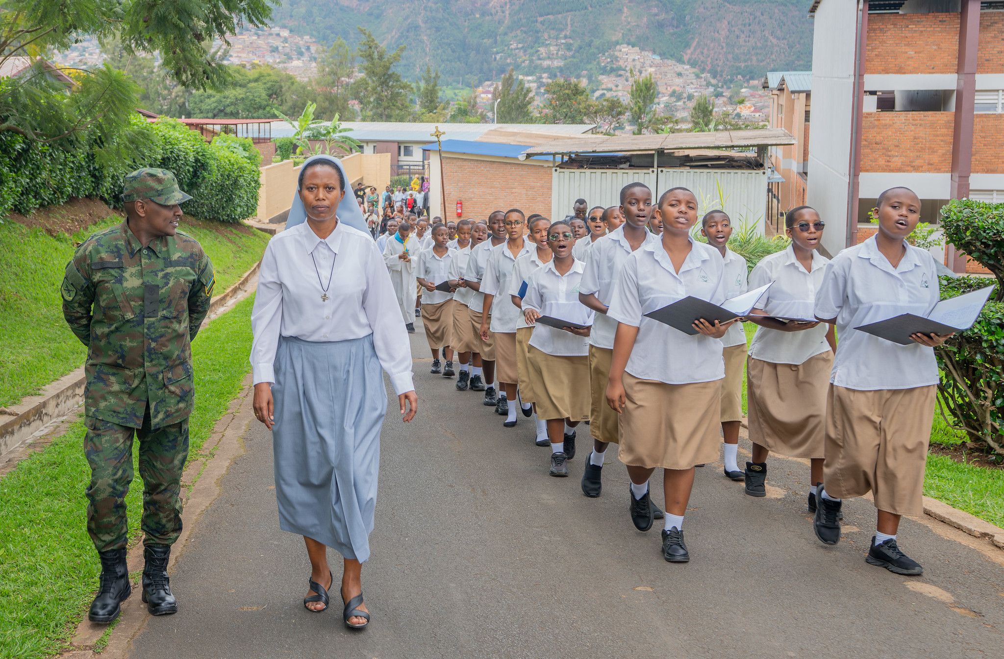 Students holding hymn books walk and sing in procession, led by a Sister and a Rwanda Defence Force officer
