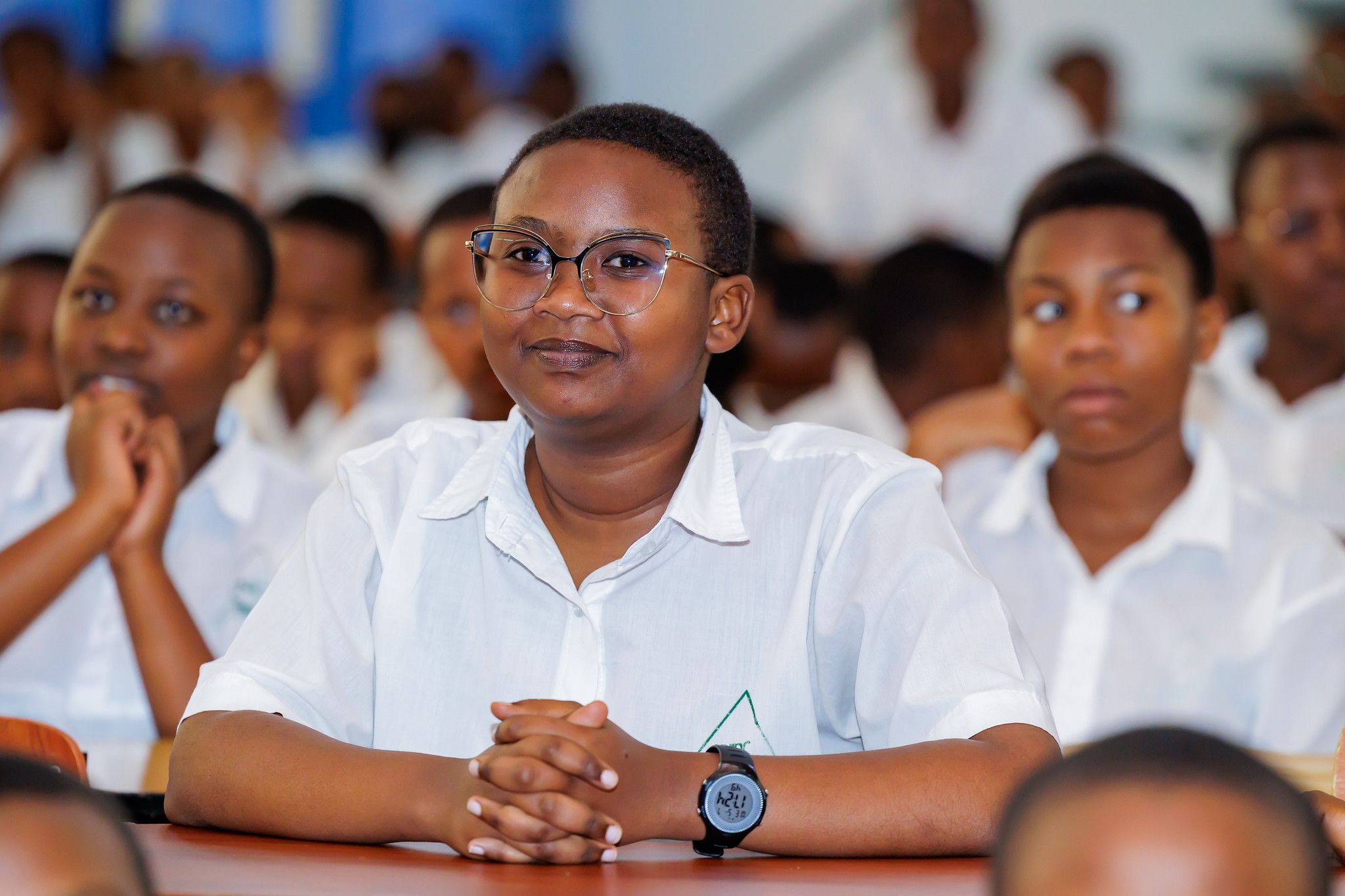 A smiling student in glasses sits with her hands folded on her desk